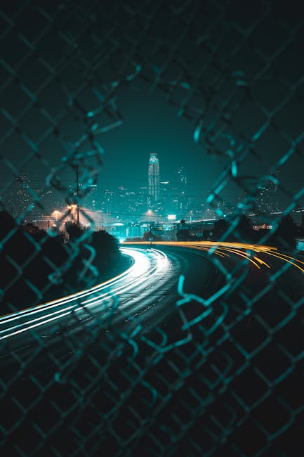 Captivating long exposure night shot of San Francisco skyline through a fence, featuring vibrant light trails.